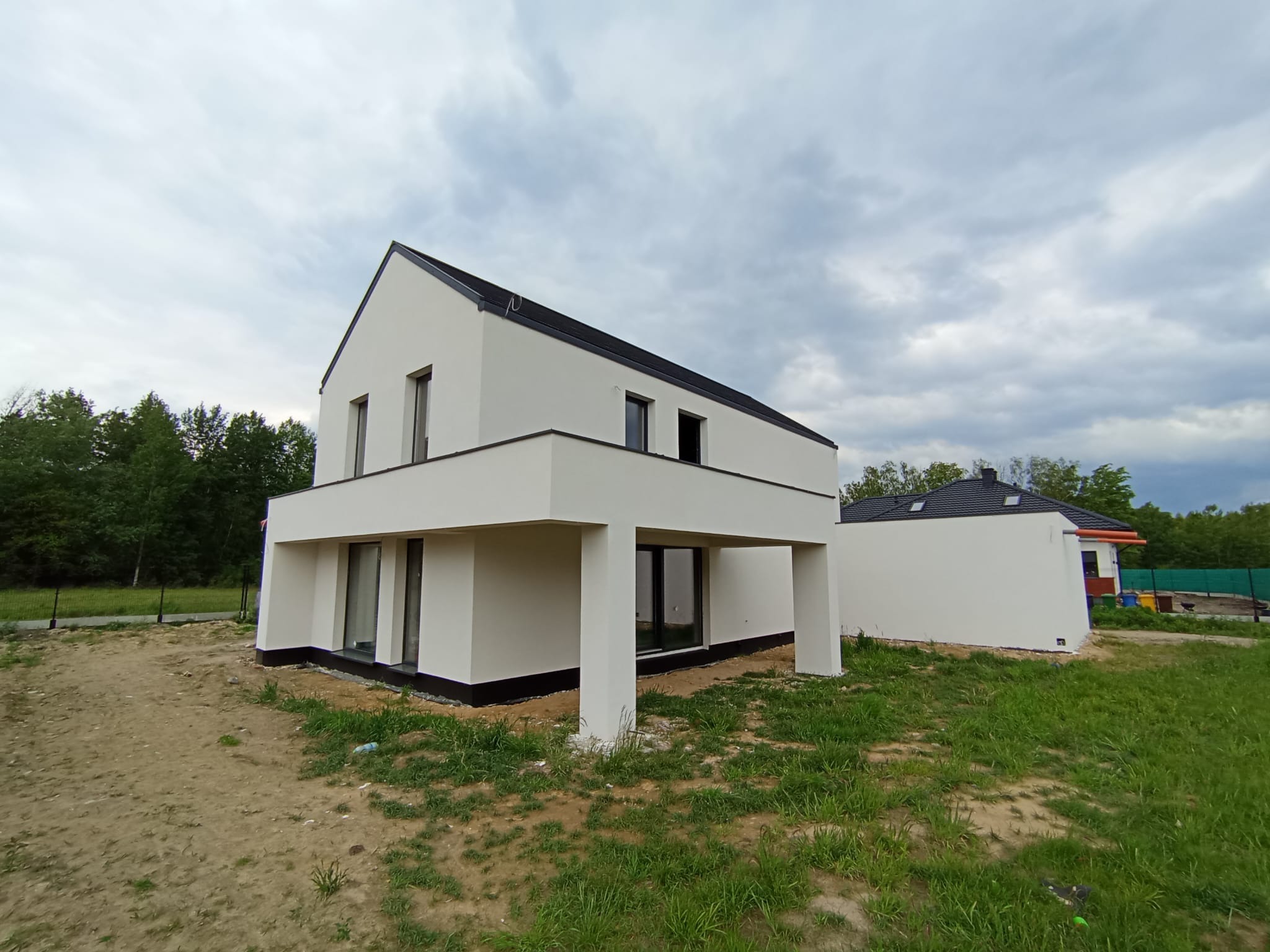 Nowoczesny, biały dom z płaskim dachem i podcieniem wspartym na kolumnach, na tle pochmurnego nieba i zieleni. Widoczny drugi budynek w tle.