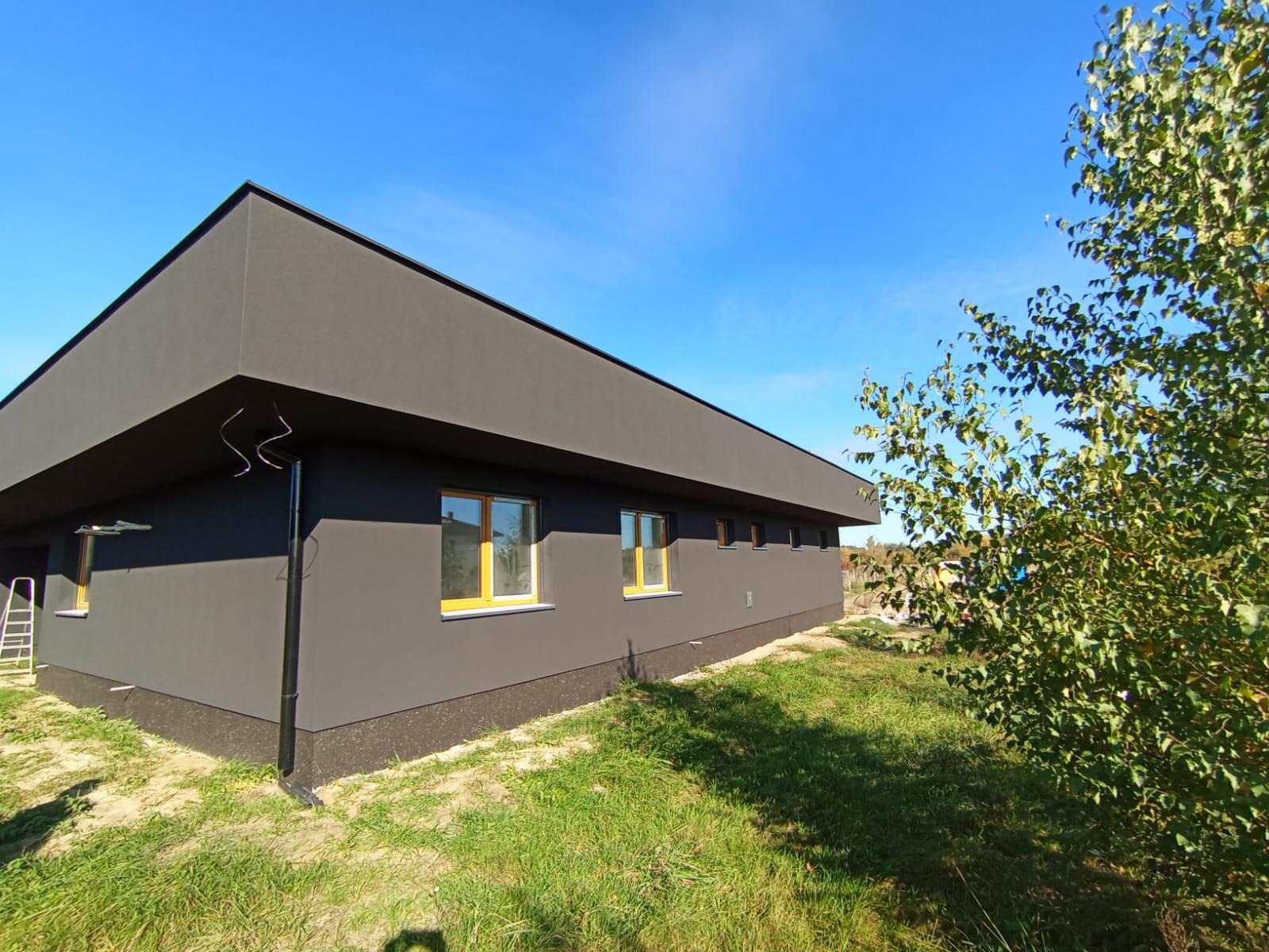 Nowoczesny, parterowy dom z ciemną elewacją i żółtymi ramami okiennymi, otoczony zielonym trawnikiem i drzewem. Widok z boku, słoneczny dzień.