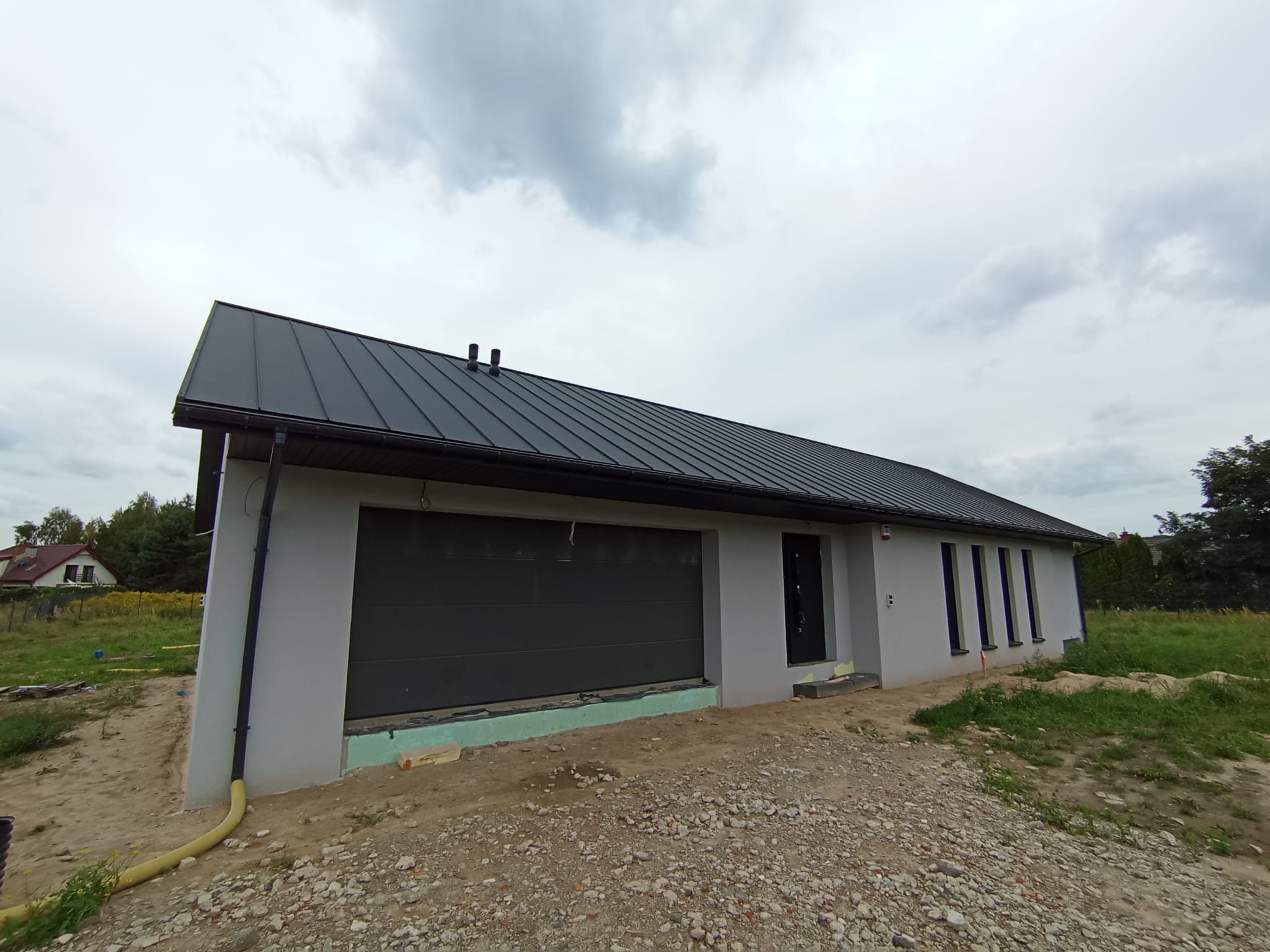 Szary dom z ciemnym dachem i garażem, widok z zewnątrz. Widoczne ocieplenie fundamentów i rury odprowadzające wodę. Dom w trakcie wykończenia.