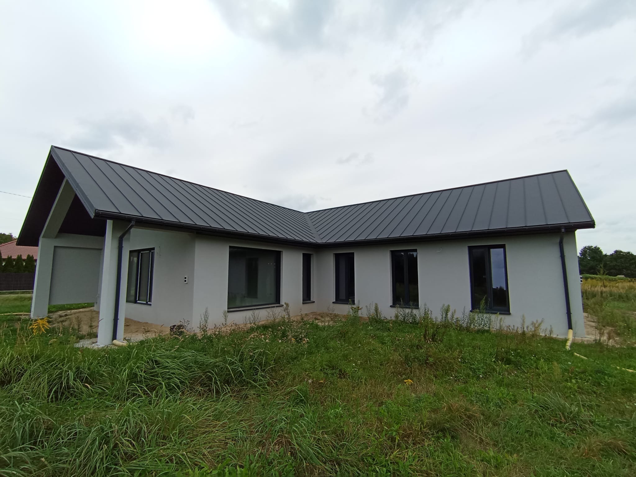 Parterowy dom z keramzytu z ciemnym dachem na tle pochmurnego nieba. Widoczne czarne okna i fragment zadaszonego tarasu. Działka zarośnięta trawą.