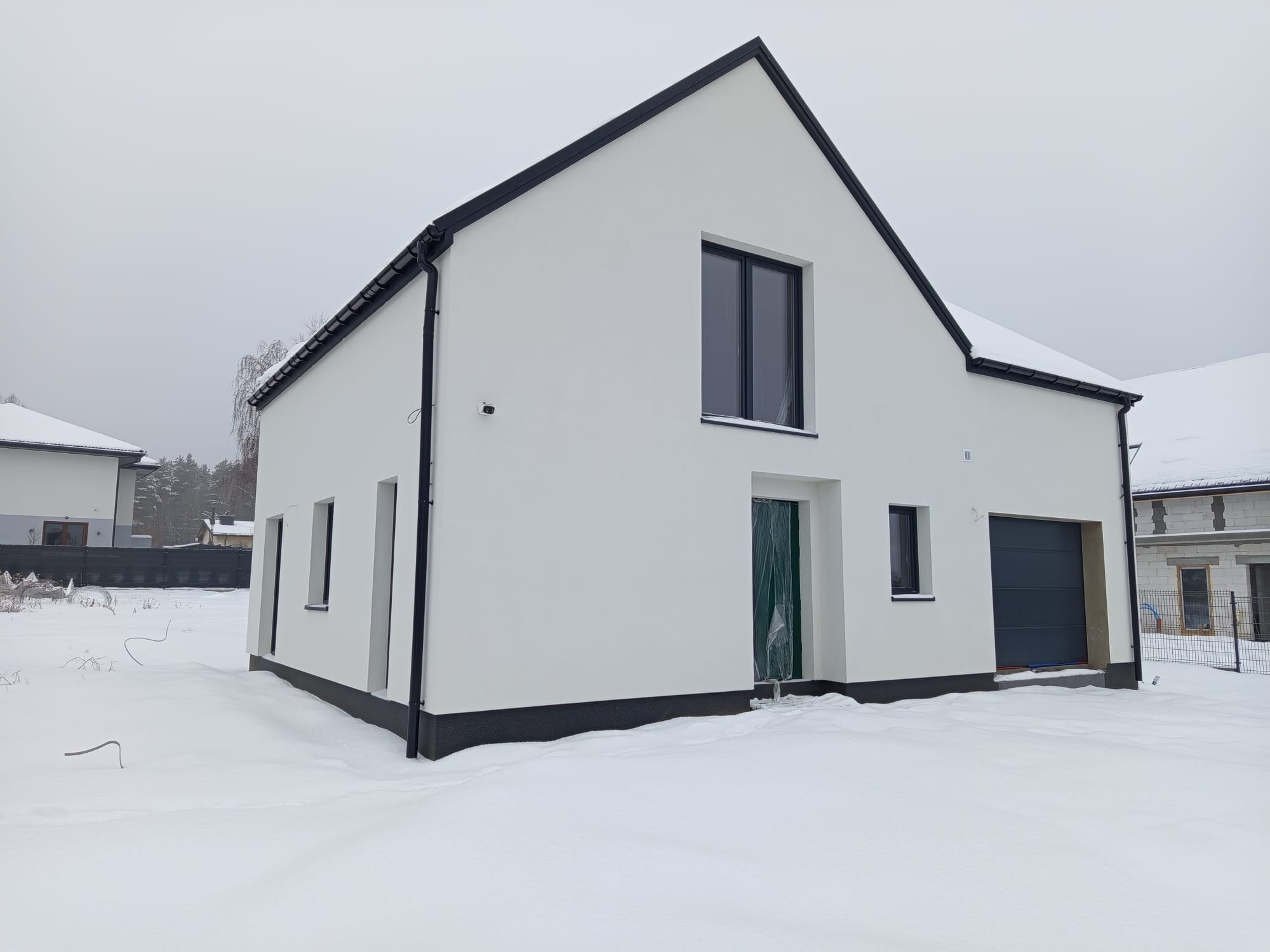 Nowoczesny, biały dom jednorodzinny z płaskim dachem i czarnymi obramowaniami okien, pokryty śniegiem, z garażem po prawej stronie.