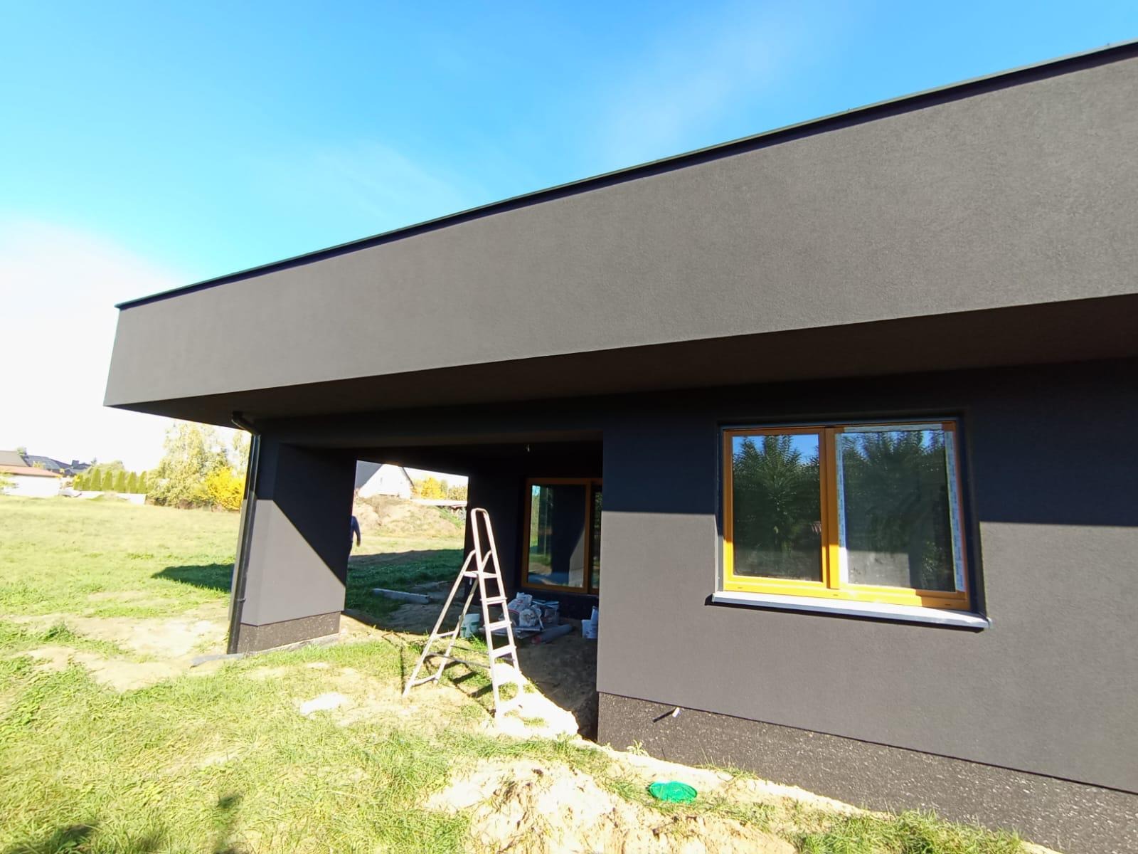 Nowoczesny dom z ciemną elewacją i żółtymi ramami okiennymi w trakcie budowy, z drabiną opartą o ścianę i widocznym trawnikiem.