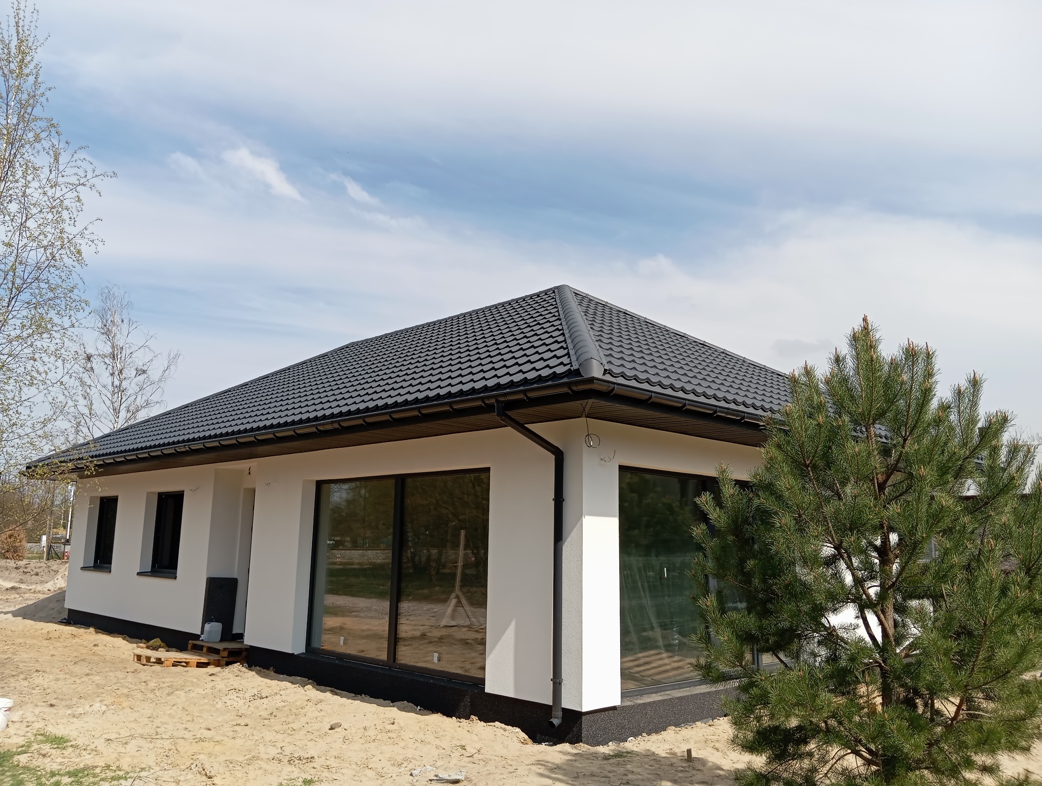 Nowoczesny, parterowy dom z ciemną dachówką i dużymi oknami, otoczony piaszczystym terenem i młodymi drzewami. Widoczne detale wykończenia elewacji.