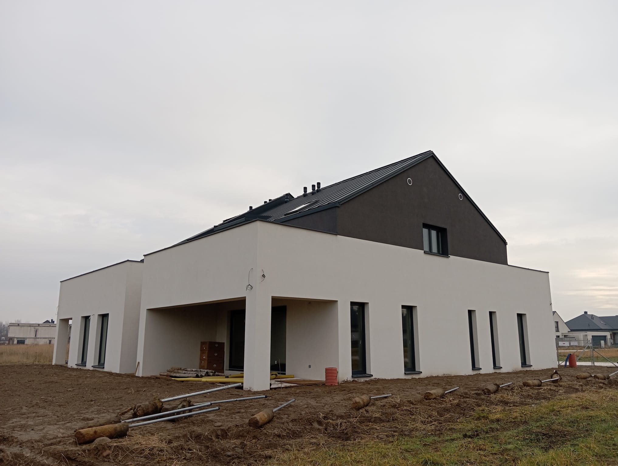 Nowoczesny, parterowy dom z płaskim dachem w trakcie budowy, z widocznymi elementami instalacji zewnętrznej i surowym otoczeniem.