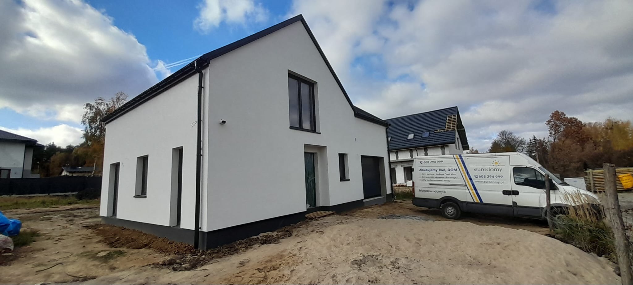 Nowoczesny dom jednorodzinny z białymi ścianami i czarnymi detalami, widoczny z zewnątrz, z samochodem dostawczym firmy budowlanej w tle.