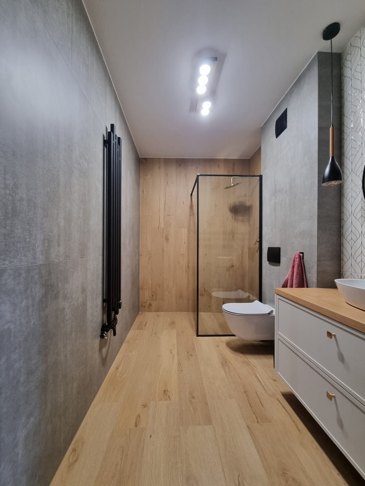 Nowoczesna łazienka po remoncie: drewnopodobne płytki, szara ściana, czarny grzejnik żeberkowy i kabina prysznicowa ze ścianką typu walk-in.