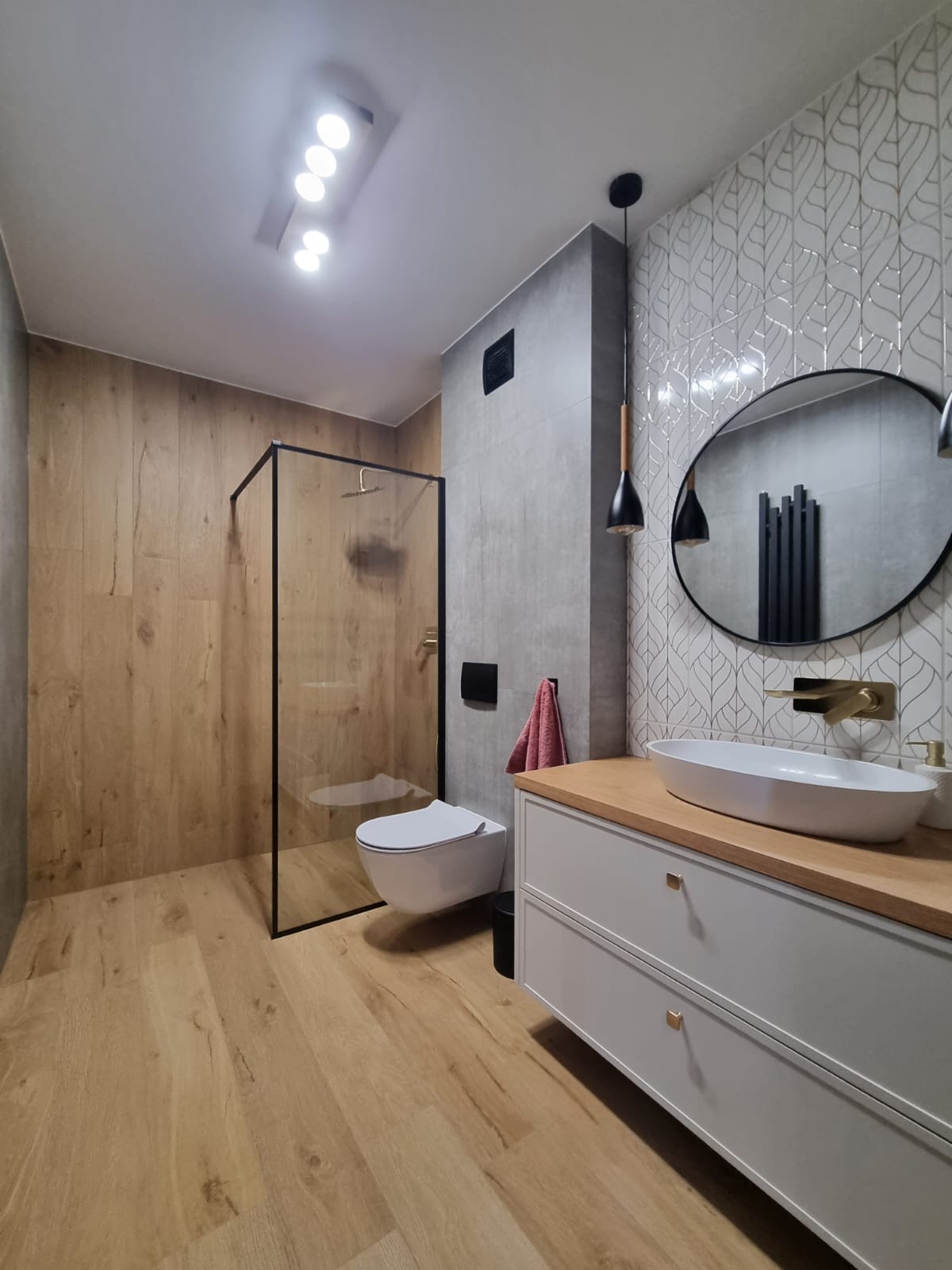 Wykończona łazienka w ciepłych barwach: drewnopodobne płytki, kabina walk-in z czarnymi ramami, biała szafka z drewnianym blatem i owalną umywalką.