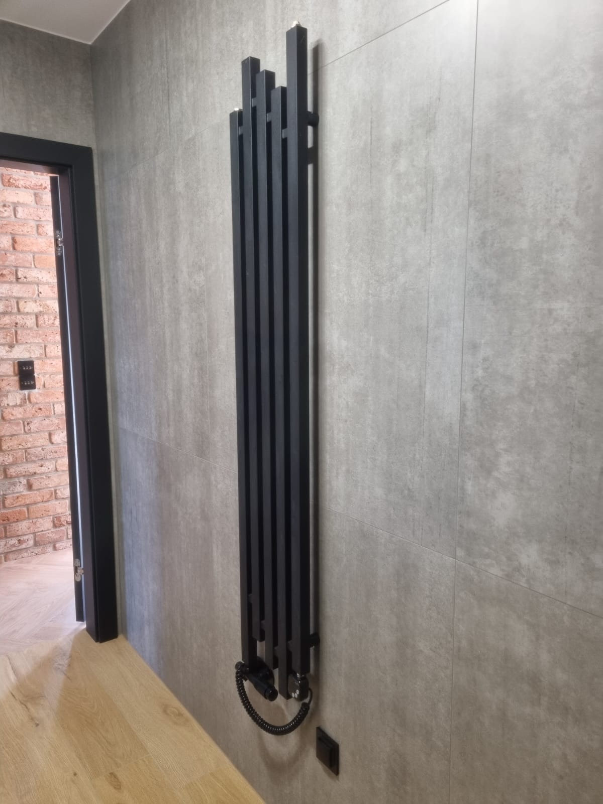 Nowoczesny grzejnik dekoracyjny, czarny, pionowy, na tle szarej ściany z płytek. Widoczny fragment podłogi i drzwi do pomieszczenia z ceglaną ścianą.