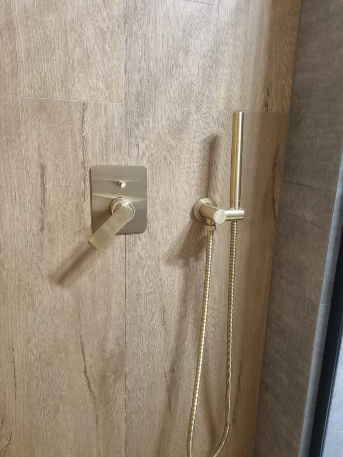 Bateria natryskowa w kolorze szczotkowanego złota, zamontowana na ścianie wyłożonej płytkami imitującymi drewno. Minimalistyczny design.