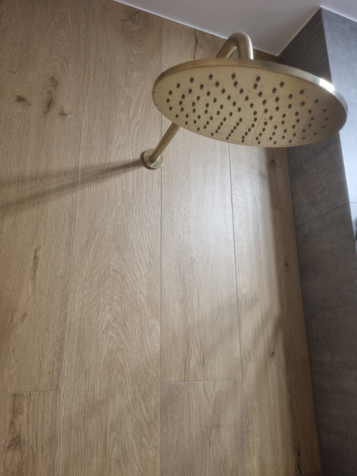 Kabina prysznicowa z deszczownicą w złotym kolorze na tle drewnopodobnych paneli ściennych i szarej ściany. Nowoczesny design łazienki po remoncie.