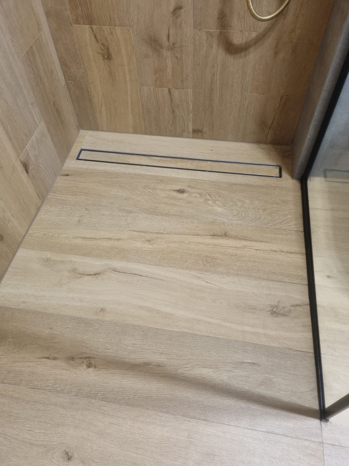 Wykończona kabina prysznicowa z odpływem liniowym i drewnopodobnymi płytkami na ścianach i podłodze, widok z góry, fragment szklanej ścianki.