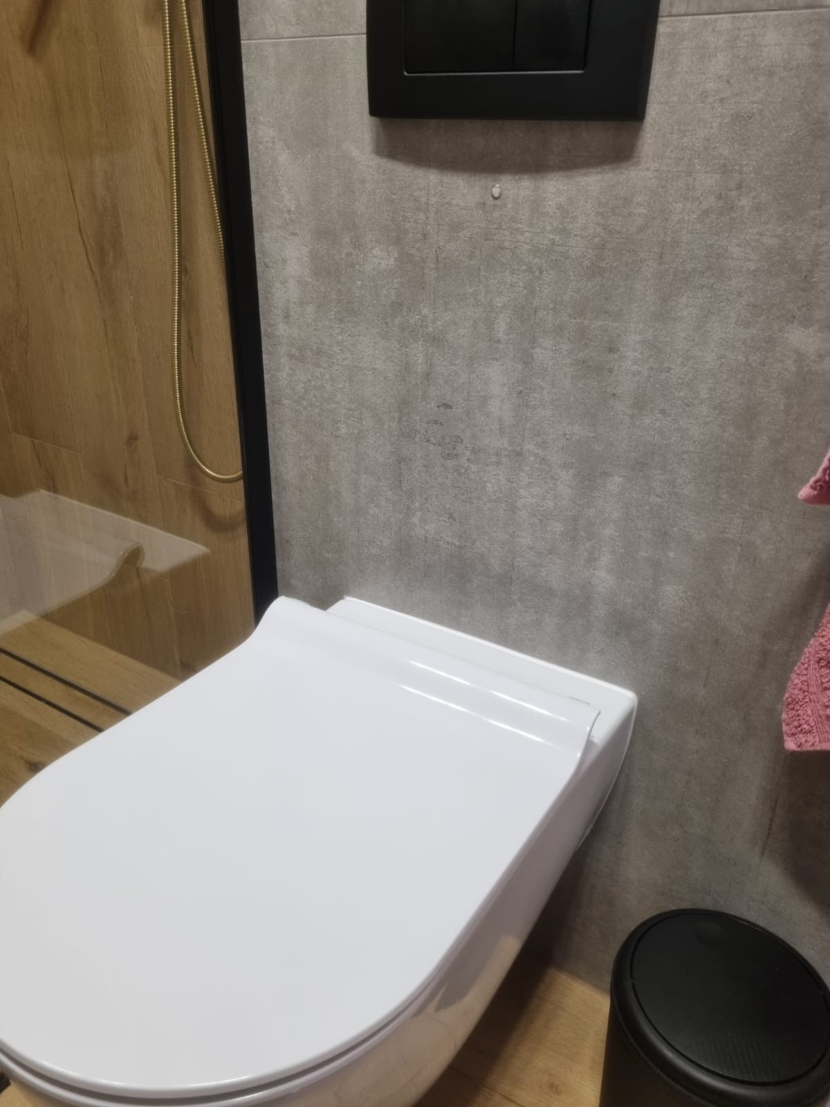 Nowoczesna, podwieszana toaleta w wykończonej łazience. Szara ściana z czarnym przyciskiem spłukiwania, drewnopodobna kabina prysznicowa z złotym wężem.