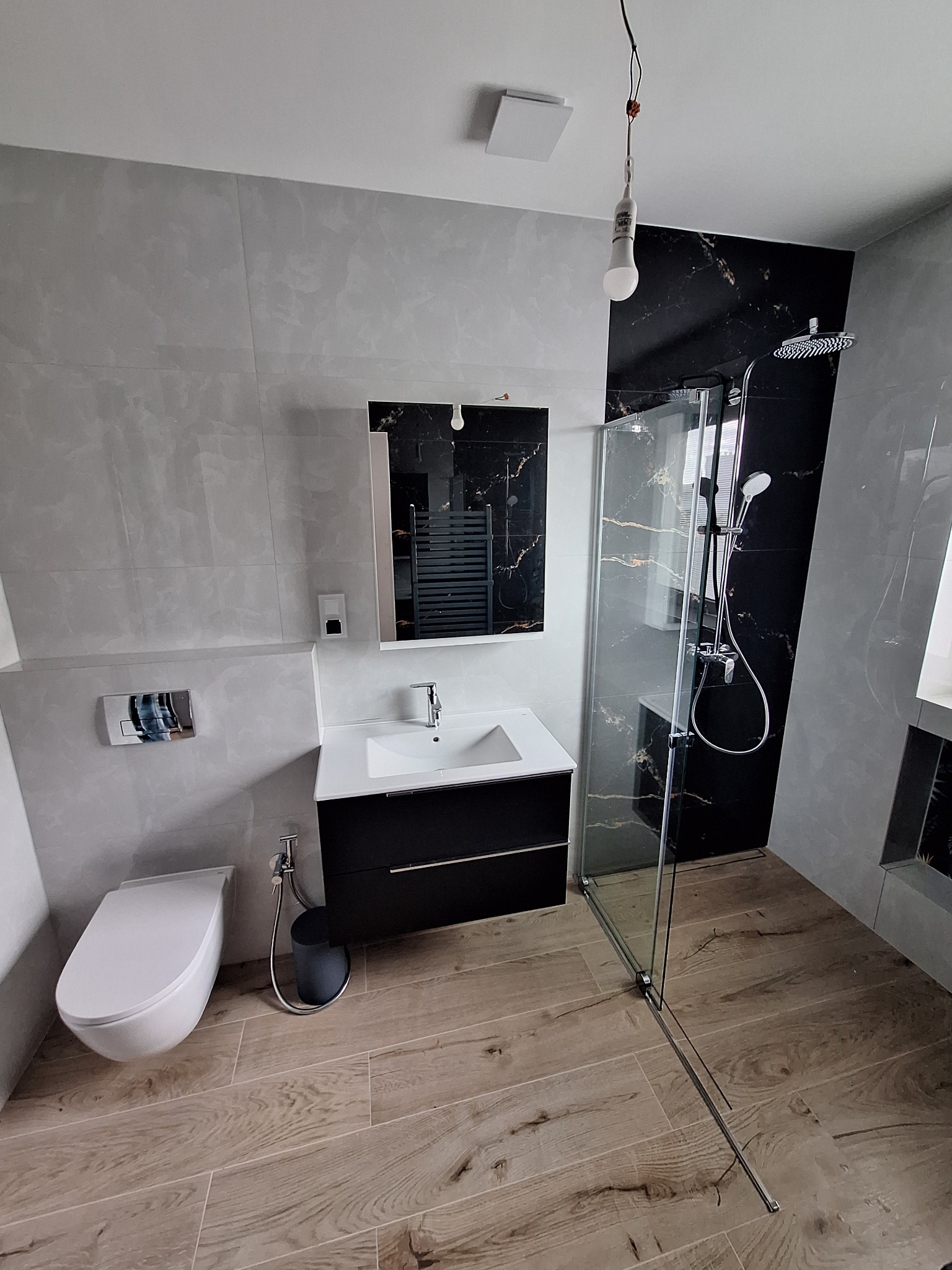 Nowoczesna łazienka po remoncie: kabina prysznicowa ze szklaną ścianką, umywalka z szafką, toaleta podwieszana, płytki imitujące marmur i drewno.