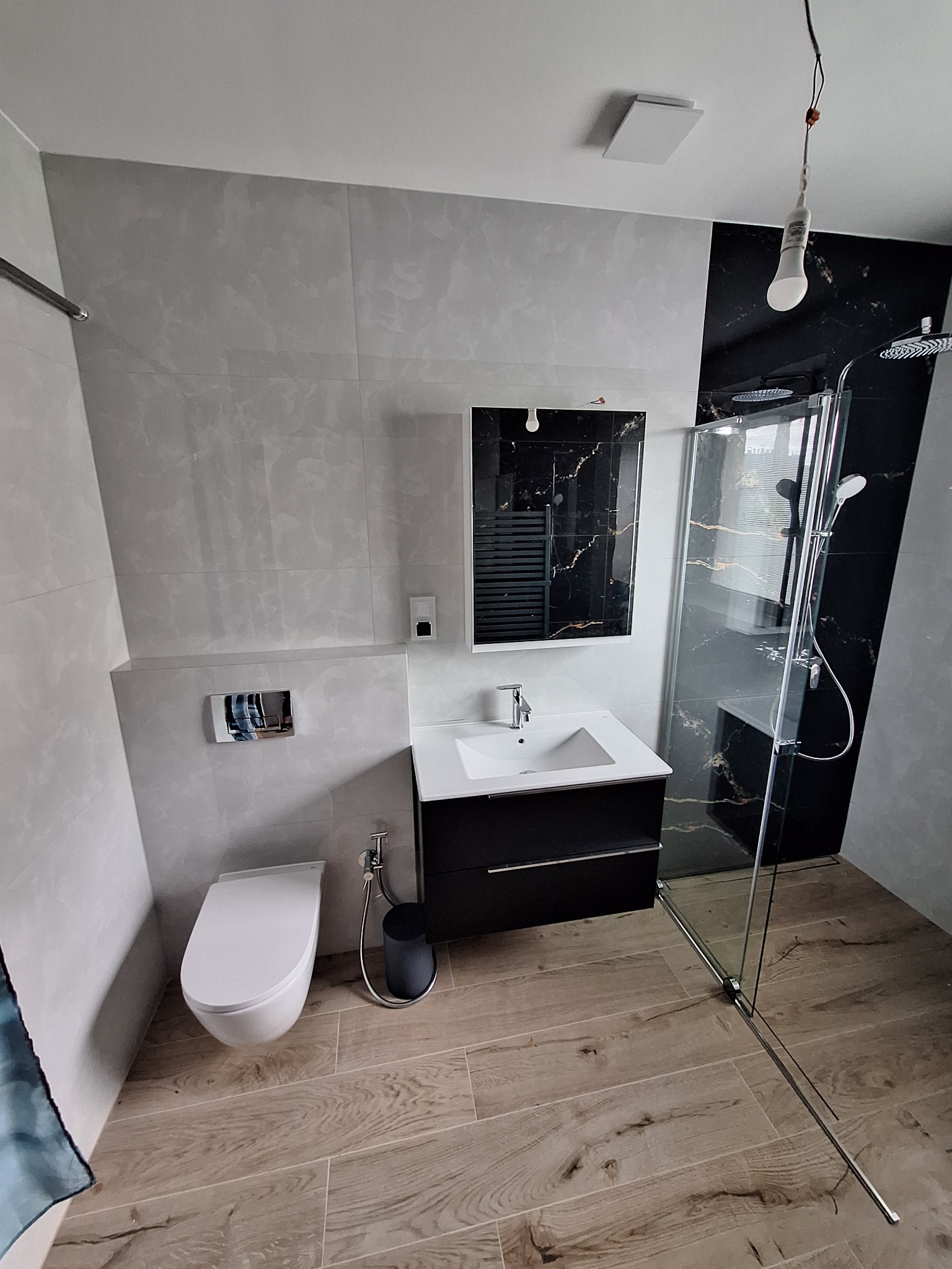 Nowoczesna łazienka po remoncie: jasne płytki imitujące marmur, drewnopodobna podłoga, czarna armatura i kabina prysznicowa z przezroczystego szkła.