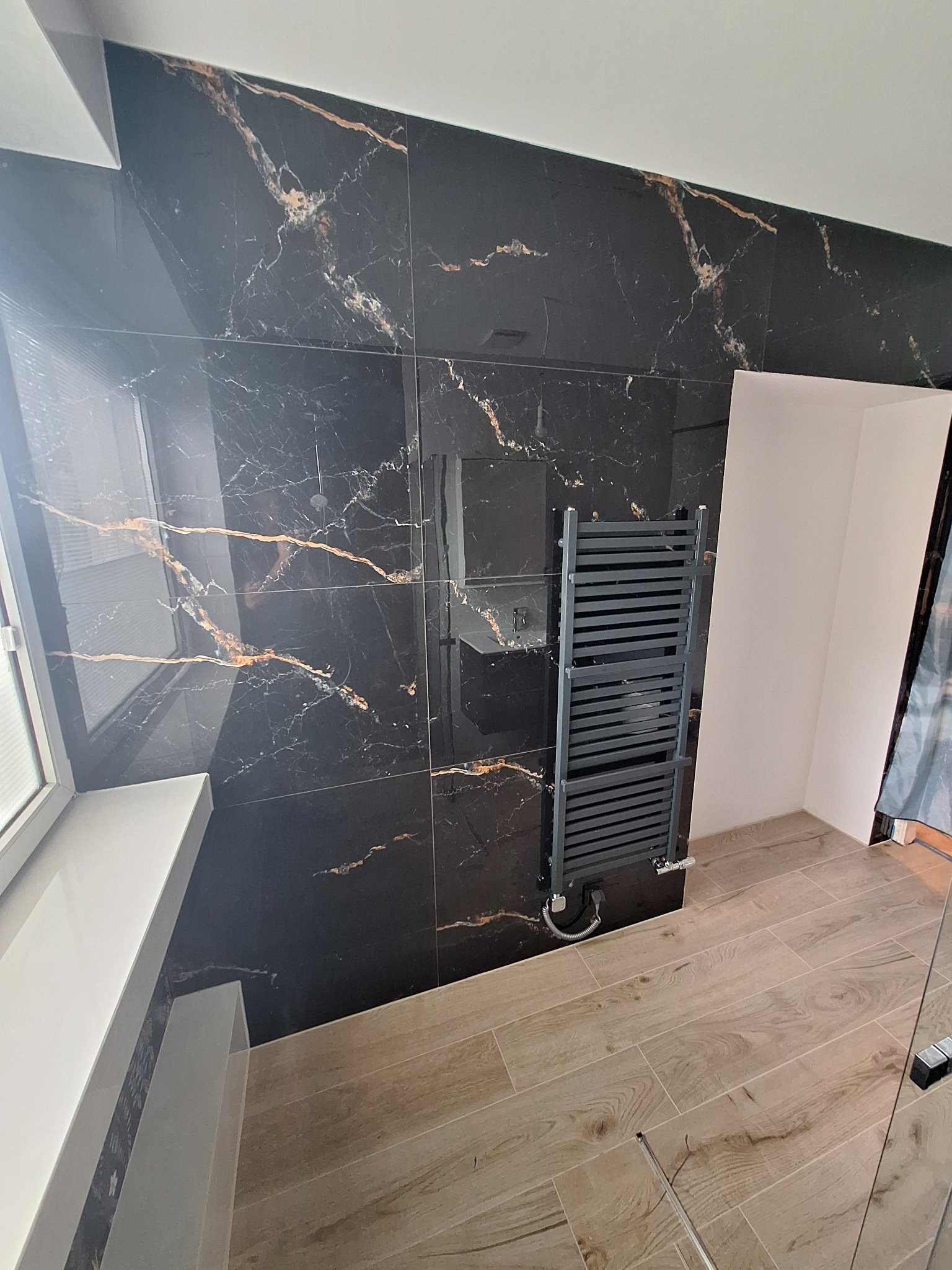 Wykończona łazienka z czarnymi płytkami imitującymi marmur i drewnopodobną podłogą. Grzejnik drabinkowy w kolorze grafitowym. Nowoczesny design.