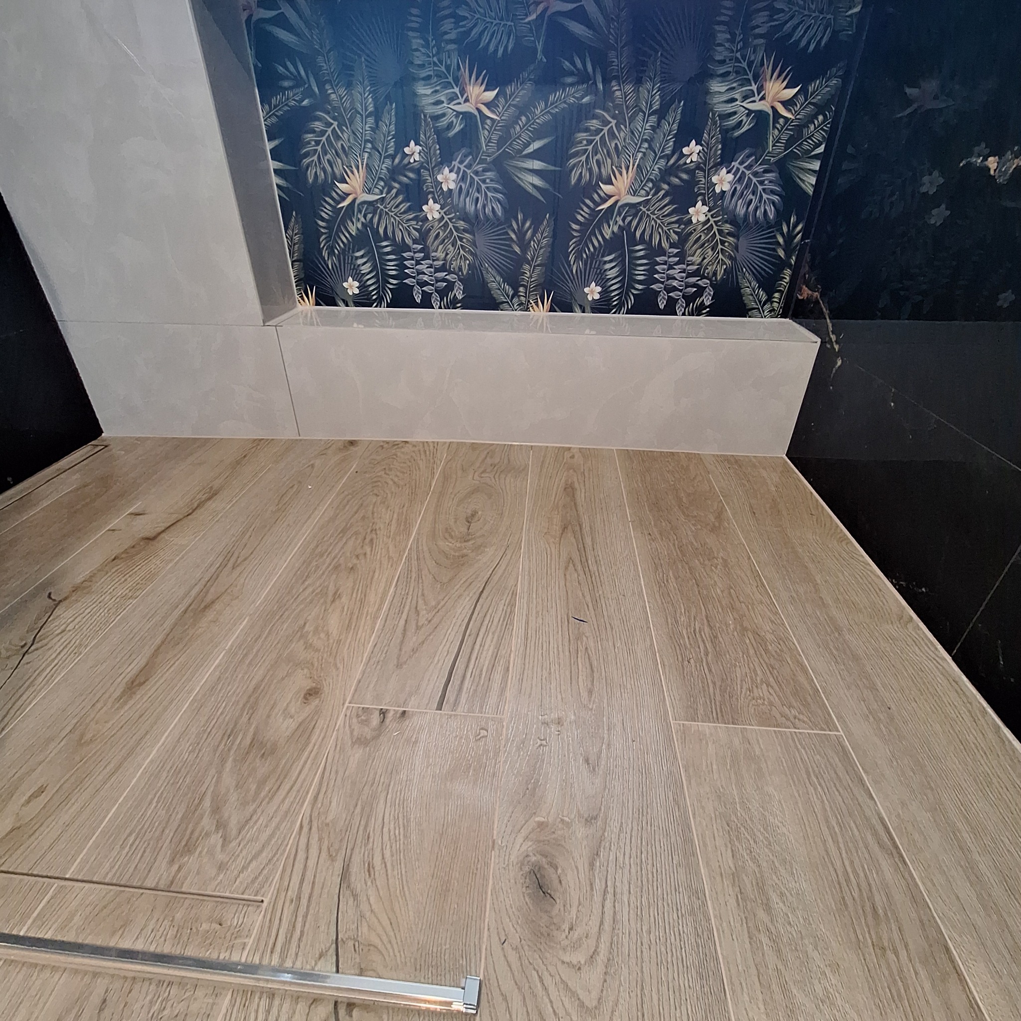 Wykończona łazienka: drewnopodobna podłoga z listwą, ściana z szarych płytek i tapeta w tropikalne liście. Nowoczesny design z dbałością o detale.