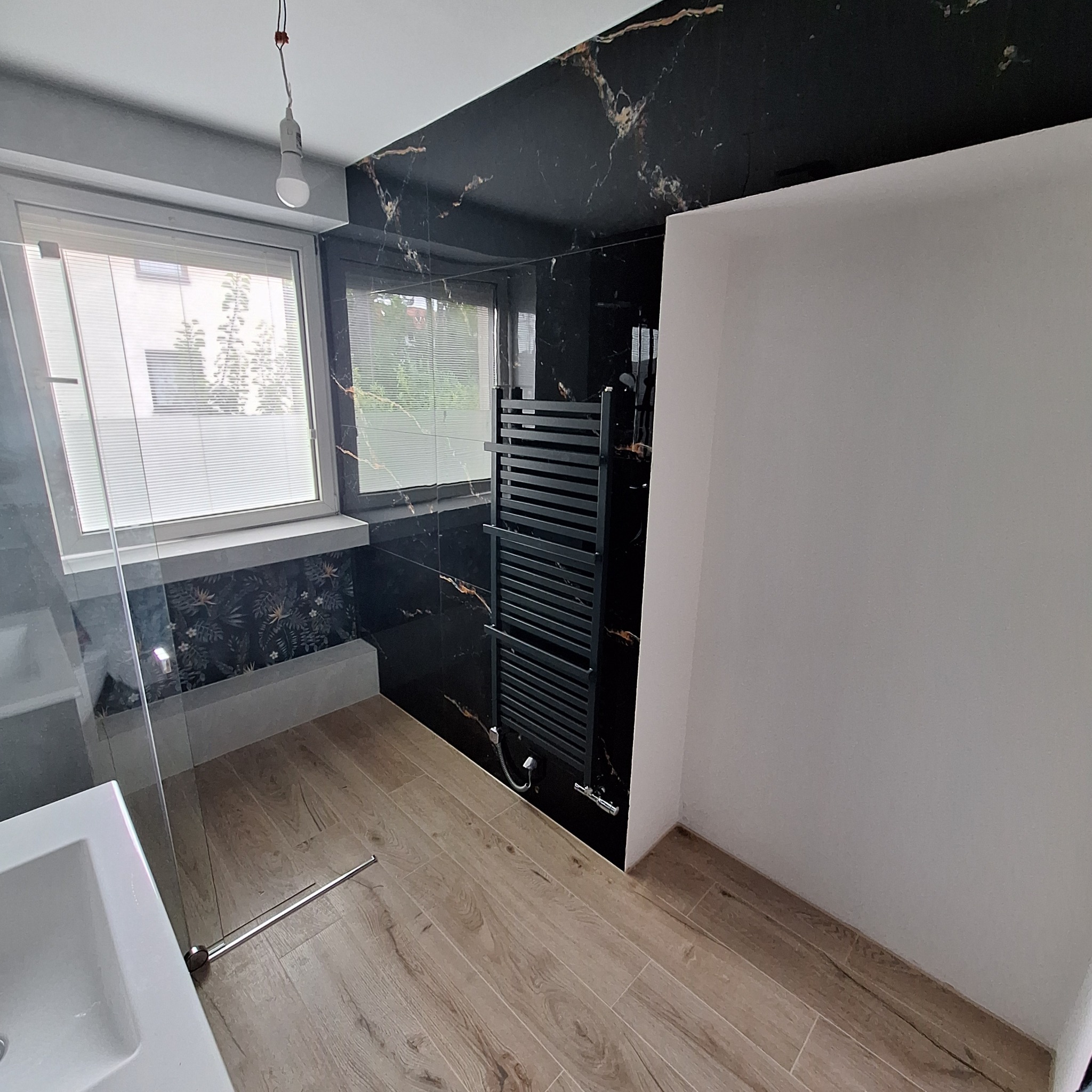 Wykończona łazienka z czarnym marmurem, drewnopodobną podłogą, grzejnikiem drabinkowym i umywalką. Widok na okno z żaluzjami.