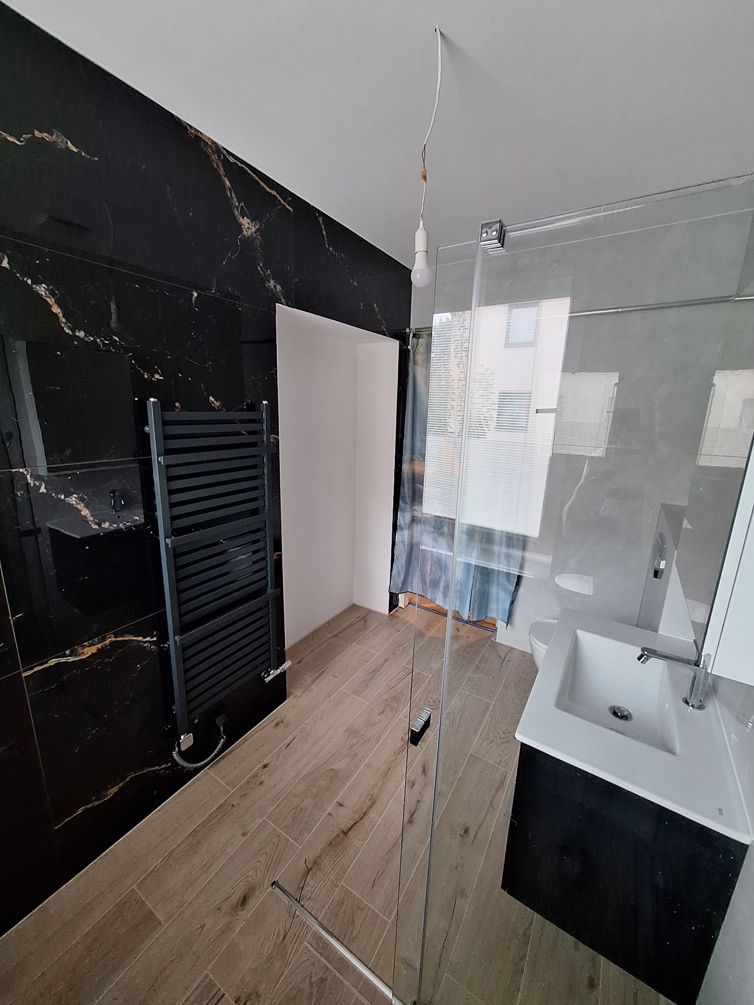 Nowoczesna łazienka po remoncie: czarne marmurowe płytki, drewnopodobna podłoga, szklana kabina prysznicowa i grzejnik drabinkowy. Minimalistyczny design.