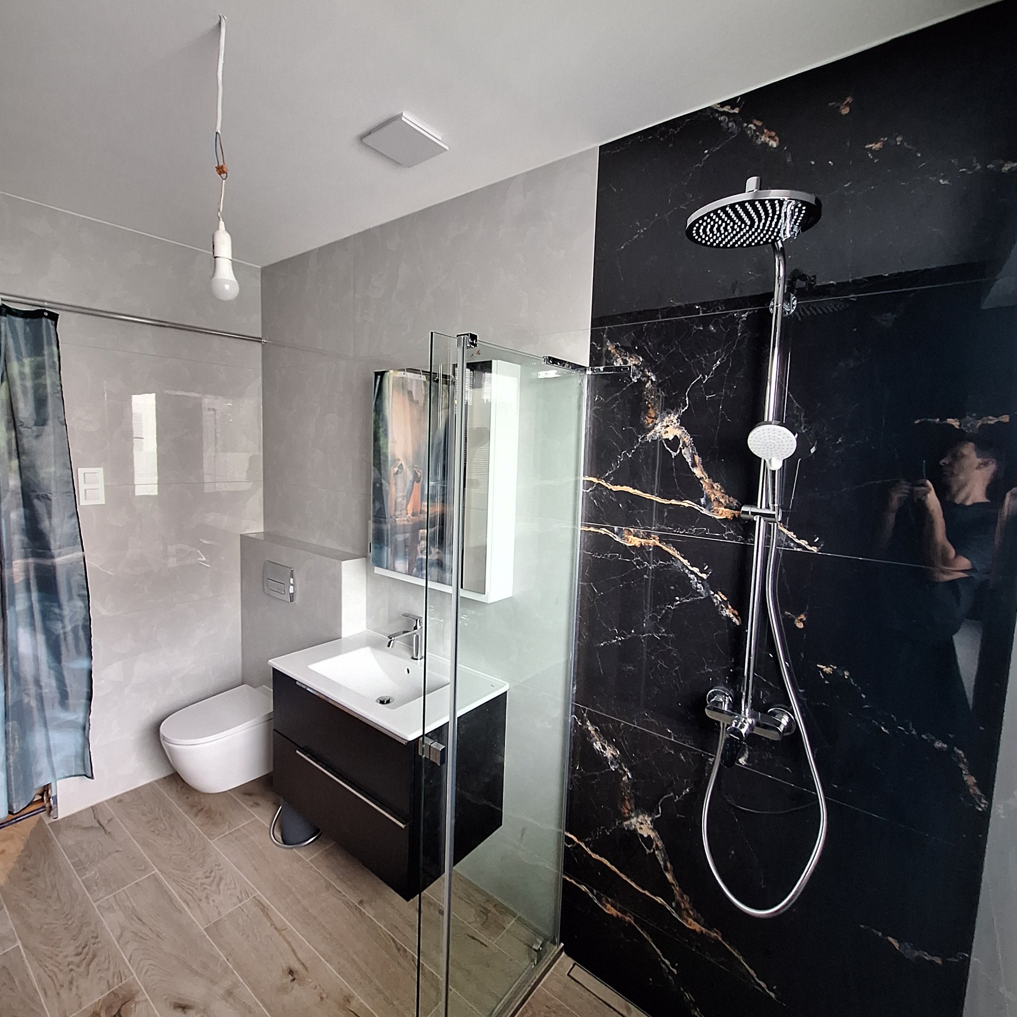 Wykończona łazienka z kabiną typu walk-in, umywalką z szafką i toaletą. Na ścianach szare i czarne płytki imitujące marmur. Widoczna niedokończona instalacja oświetleniowa.