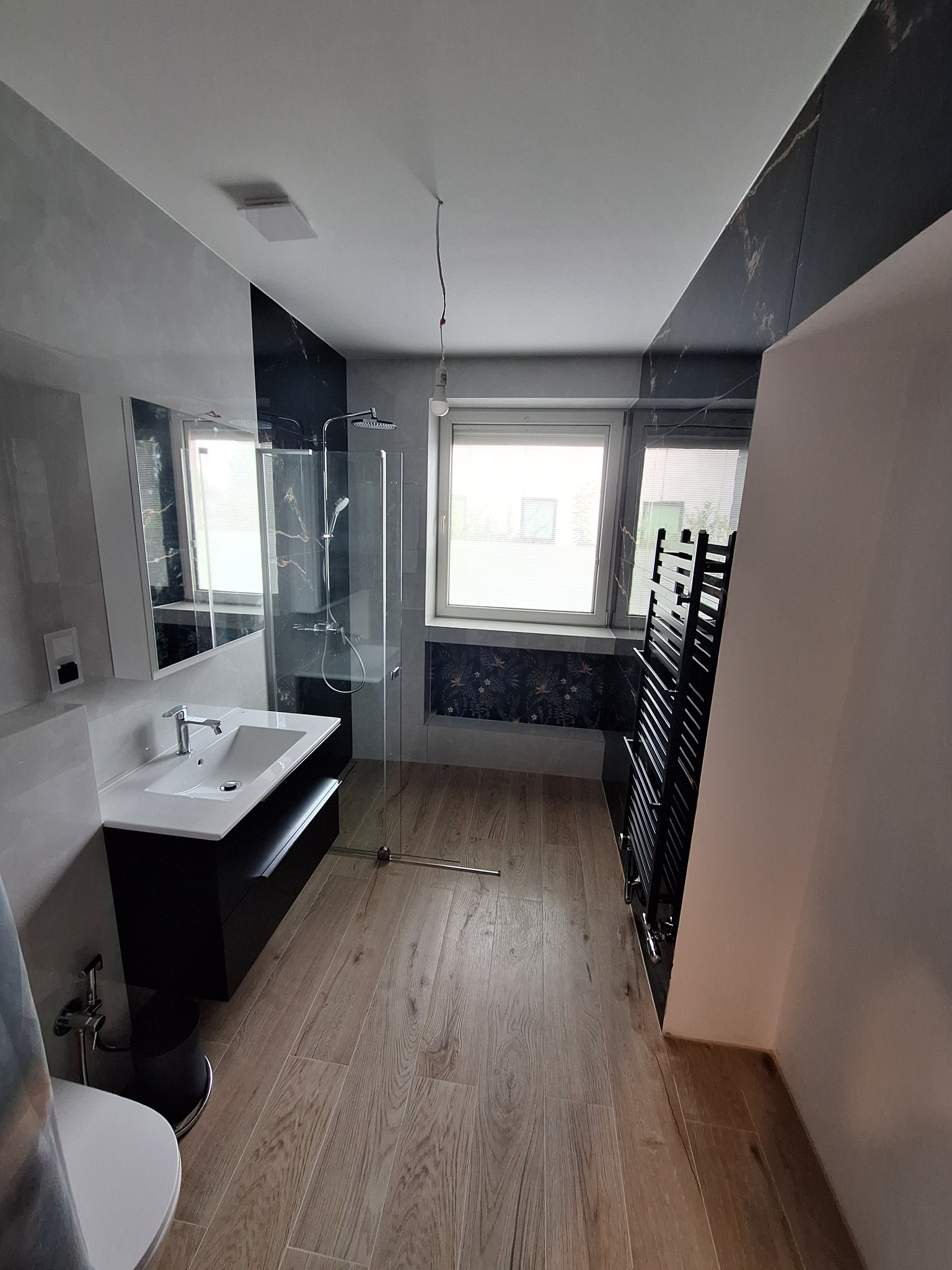 Nowoczesna łazienka z kabiną prysznicową typu walk-in, czarną szafką pod umywalką i grzejnikiem drabinkowym. Jasne drewnopodobne płytki na podłodze.