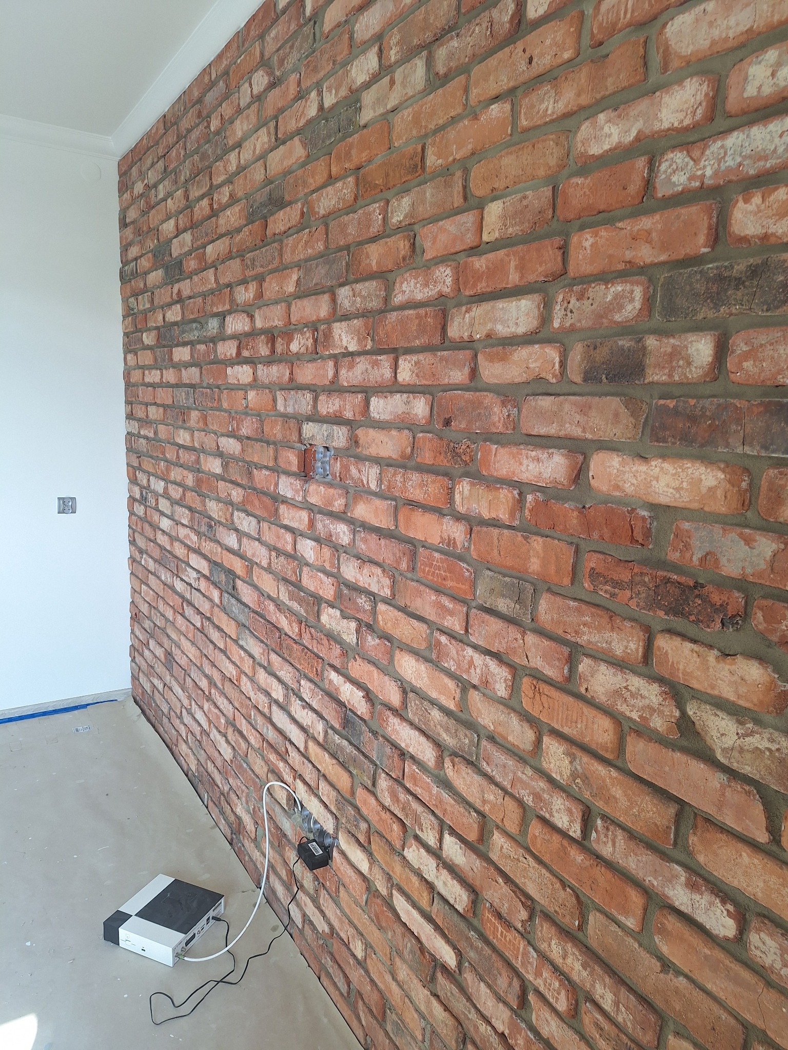 Ściana z czerwonej cegły z widoczną instalacją elektryczną. Surowy, industrialny styl wykończenia wnętrza. Widoczny fragment podłogi i listwa przypodłogowa.