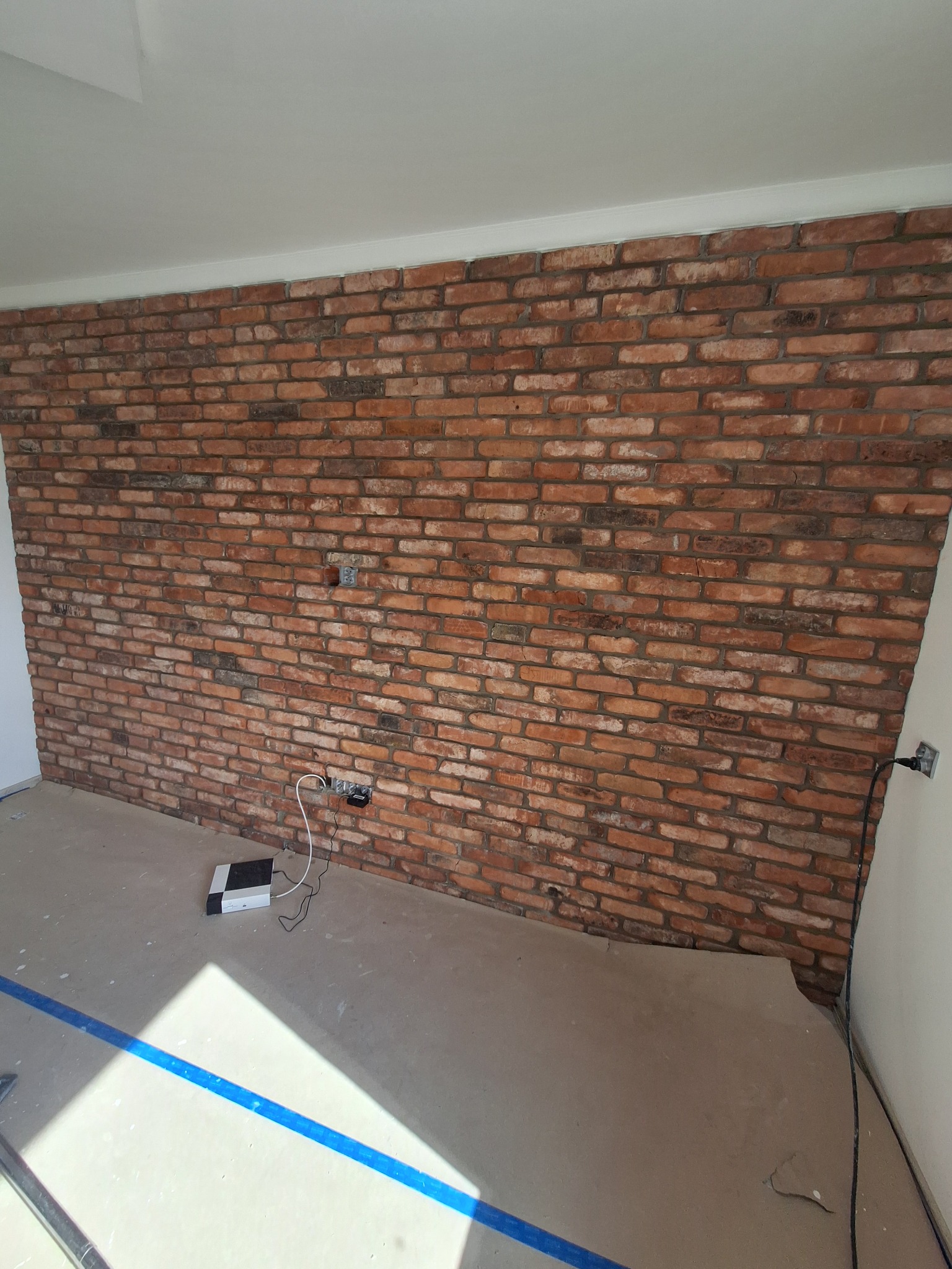 Ściana z cegły klinkierowej w pokoju, widoczne gniazdka elektryczne i sprzęt elektroniczny na podłodze, niebieska taśma malarska. Wykończenie wnętrza w stylu industrialnym.