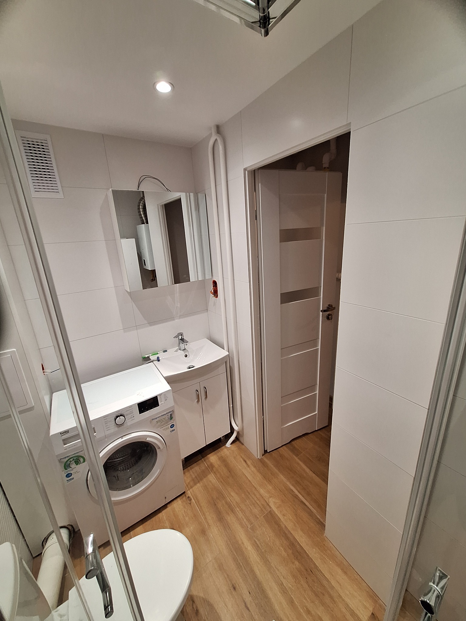 Biała łazienka z pralką, umywalką i lustrem. Na ścianach płytki, na podłodze drewnopodobne panele. Widoczny fragment kabiny prysznicowej i drzwi wejściowe.