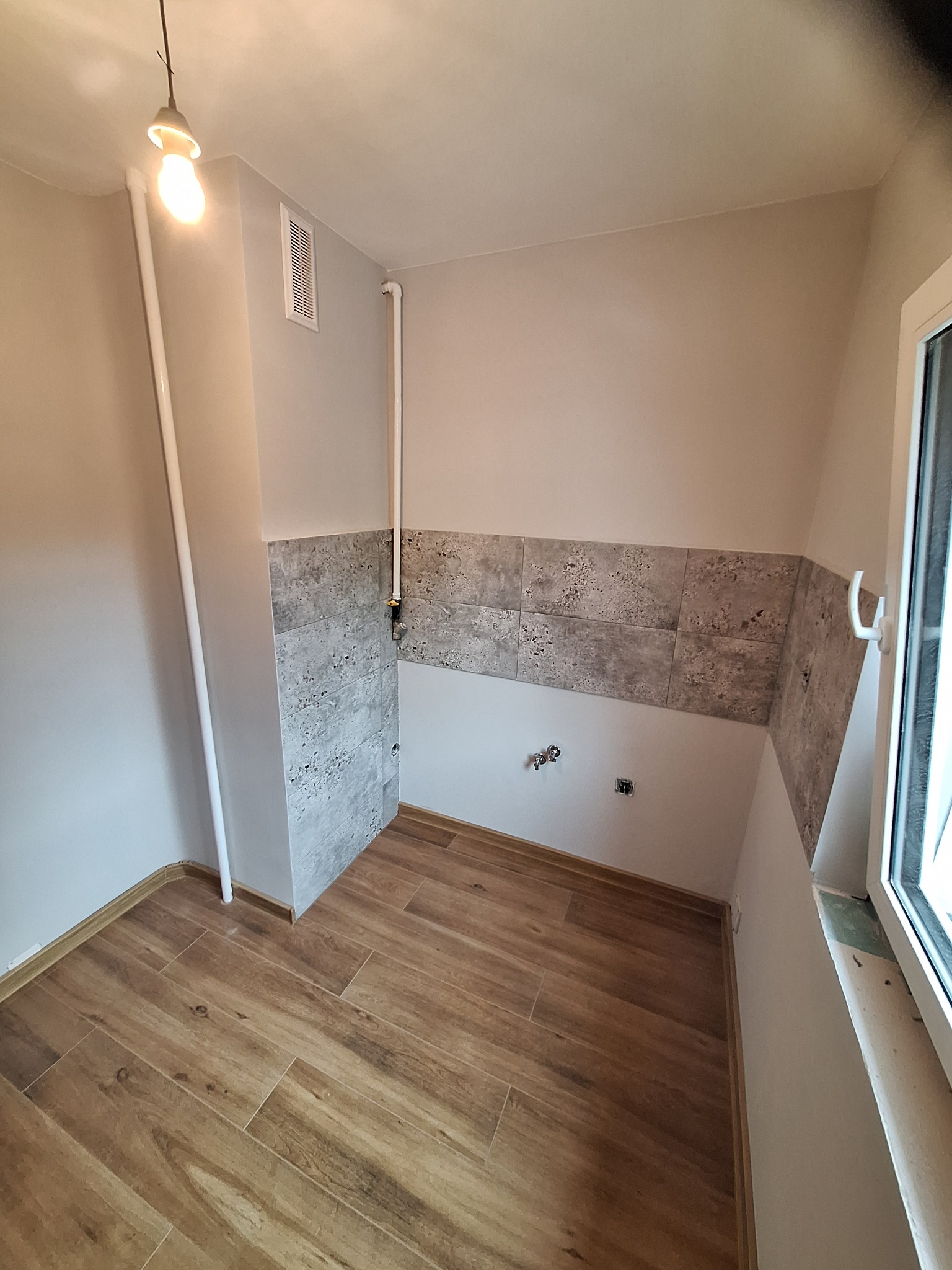 Wykończona łazienka w Częstochowie z drewnopodobną podłogą i szarymi płytkami na ścianach, widoczne instalacje wodne i okno.