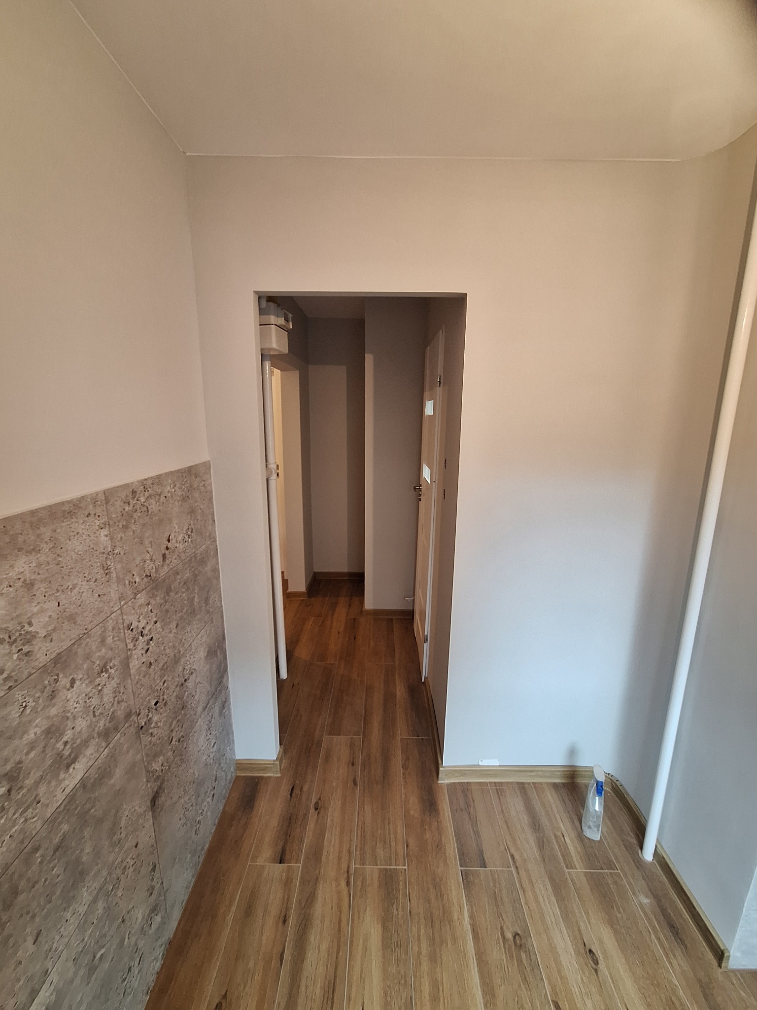 Wykończony korytarz z drewnopodobną podłogą i fragmentem ściany z dekoracyjną okładziną. Widoczne drzwi w głębi i biała rura przy ścianie.