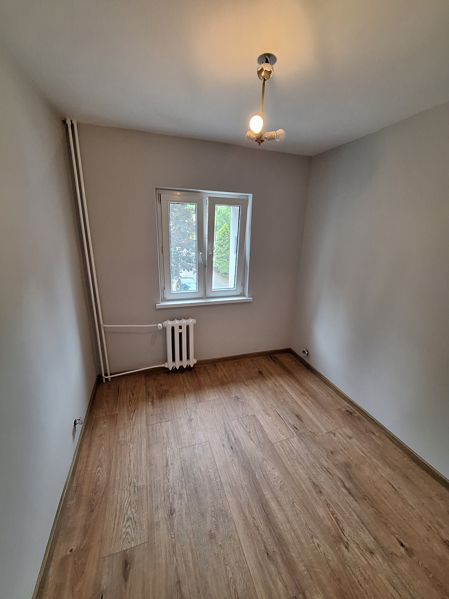 Pusty pokój po remoncie: drewniana podłoga, białe ściany, okno z widokiem na zieleń. Minimalistyczna lampa sufitowa. Grzejnik pod oknem.