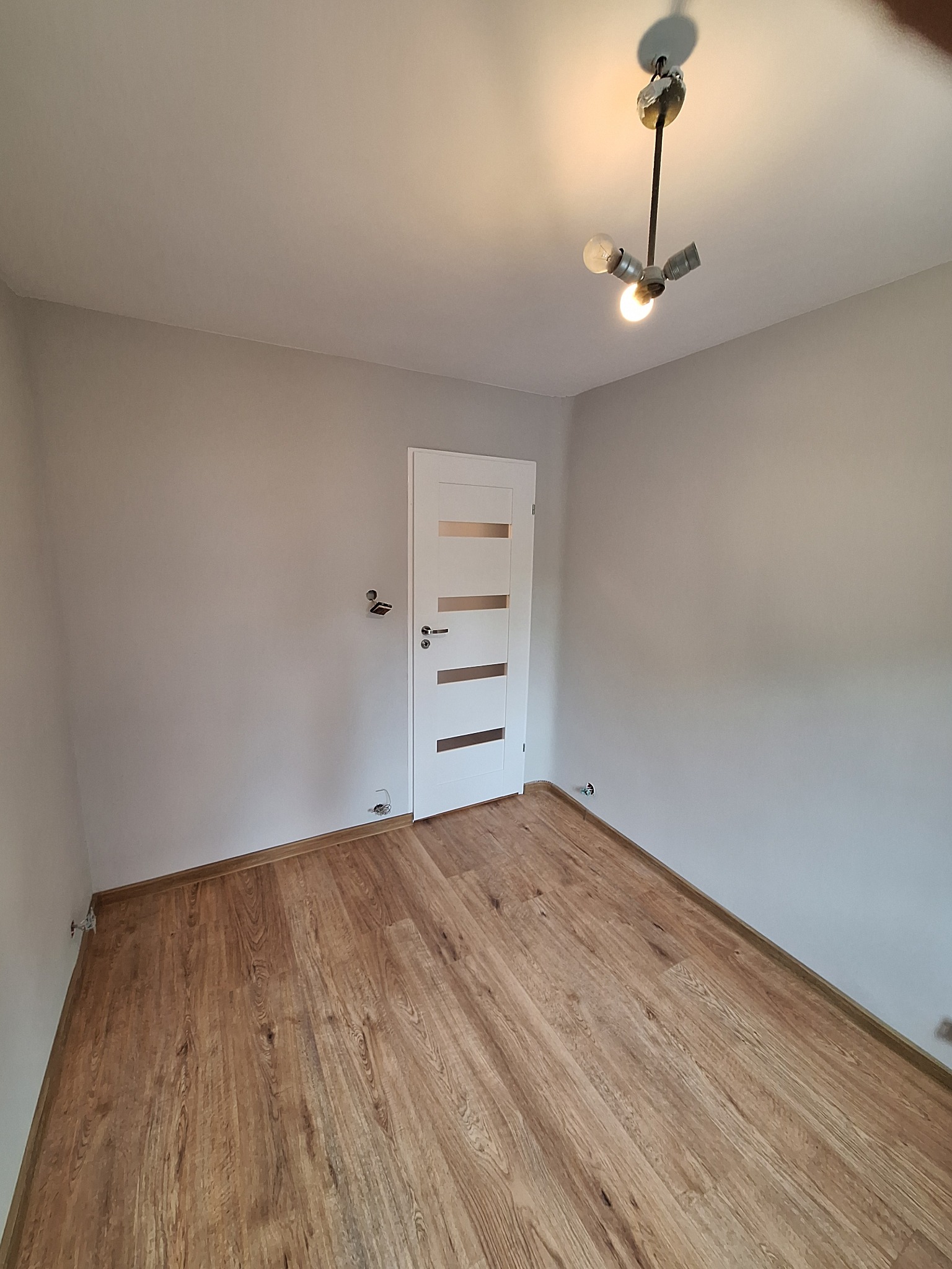 Pokój po remoncie: parkiet drewniany, białe ściany i drzwi z przeszkleniami. Widoczne niedokończone przyłącza elektryczne. Lampa sufitowa z trzema żarówkami.