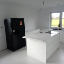 Nowoczesna, minimalistyczna kuchnia z wyspą w Ostrzeszowie, z czarną lodówką side-by-side i widokiem na zielone pola za oknem.