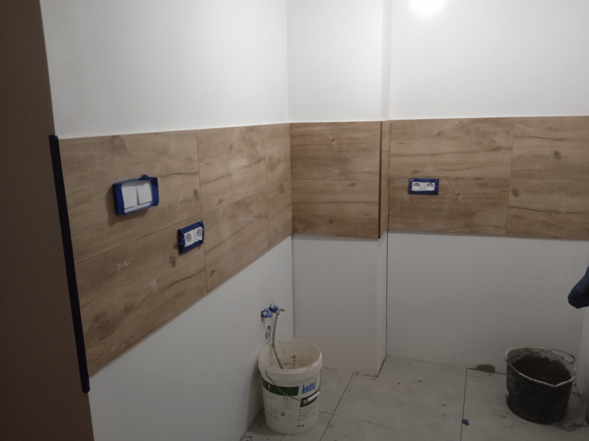 Ściana w trakcie układania płytek imitujących drewno w Poznaniu, widoczne puszki elektryczne zabezpieczone taśmą, wiadro z klejem Knauf i zaprawa w wiadrze.