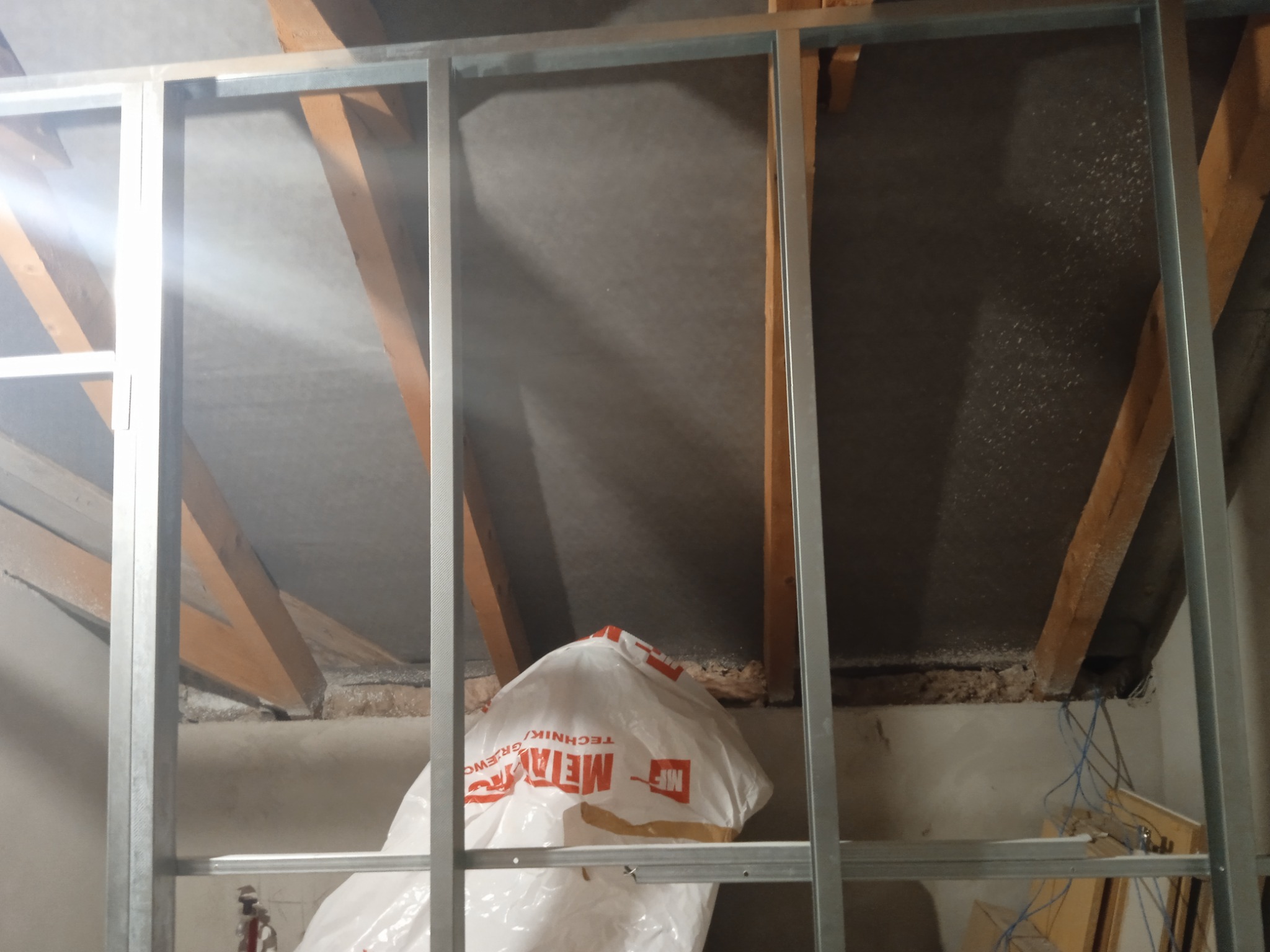 Metalowa konstrukcja szkieletowa pod płytę gipsowo-kartonową w trakcie montażu na poddaszu, widoczna izolacja i worek z materiałami budowlanymi.