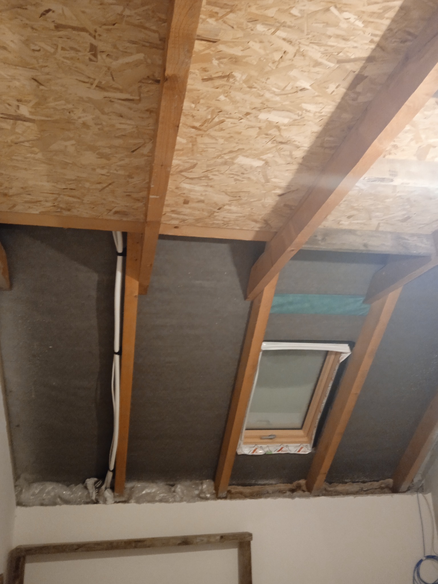 Poddasze w trakcie adaptacji: widoczne drewniane belki konstrukcyjne, płyty OSB na suficie, szara wełna mineralna między krokwiami, okno dachowe z folią ochronną, instalacja elektryczna...