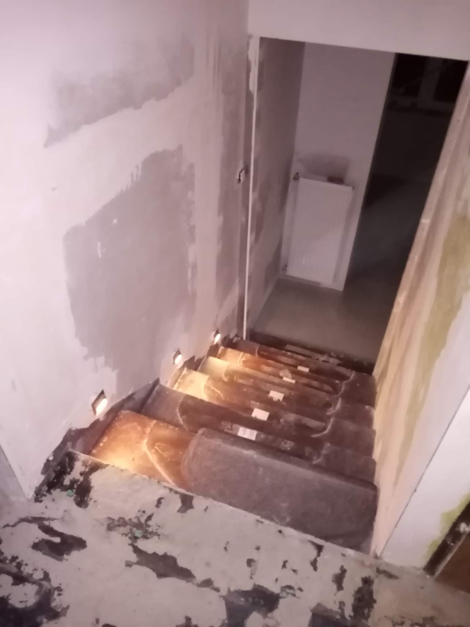 Schody w trakcie remontu z widocznymi śladami po kleju na podłodze i niedokończonymi ścianami, z małymi lampkami LED wbudowanymi w stopnie.