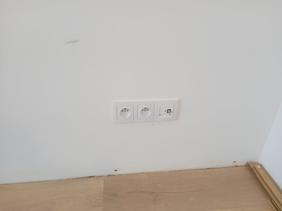 Zamontowane, potrójne gniazdko elektryczne i gniazdo antenowe w białej ramce na świeżo pomalowanej ścianie, widoczne ślady po pracach wykończeniowych na podłodze.
