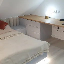 Pokój na poddaszu ze skośnym sufitem: łóżko z wezgłowiem w odcieniach beżu i pomarańczowymi poduszkami oraz biurko z blatem z jasnego drewna, pod którym znajdują się białe szuflady i szafki...