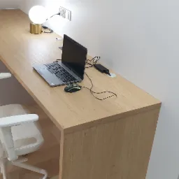 Długi, drewniany blat biurka z laptopem i myszką, zamontowany przy ścianie, z ergonomicznym krzesłem obok. Widoczne gniazdka elektryczne i lampa kulista na blacie. Minimalistyczne wnętrze.