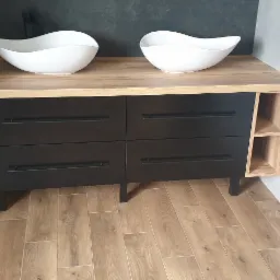 Nowoczesna szafka łazienkowa z blatem imitującym drewno, czarnymi frontami szuflad i dwoma białymi, asymetrycznymi umywalkami nablatowymi.