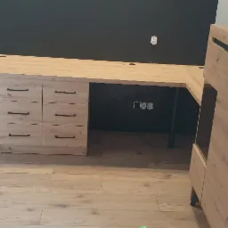 Jasny, narożny blat biurka z sześcioma szufladami i szafką, wykonany z drewna z widocznym usłojeniem, na tle czarnej ściany z gniazdkami elektrycznymi, na podłodze panele drewniane.