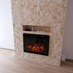 Waj-Bud Ogólno Budowlane - Ściana z dekoracyjnej, jasnej cegły z wnęką, w której umieszczony jest elektryczny kominek z efektem płomieni i imitacją drewna, na podłodze panele w odcieniach beżu.