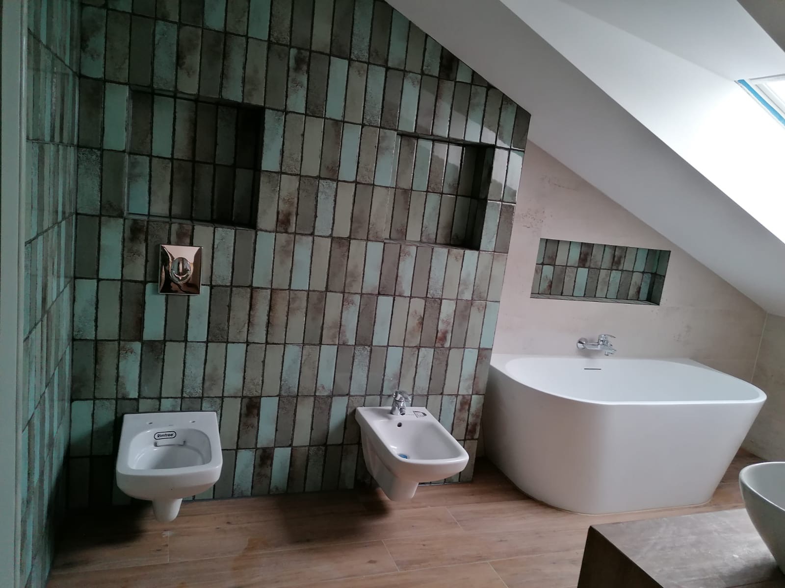 Nowoczesna łazienka na poddaszu z wanną wolnostojącą, bidetem i toaletą podwieszaną, wykończona pionowymi, ceramicznymi płytkami w odcieniach zieleni i beżu z wnękami w ścianie.