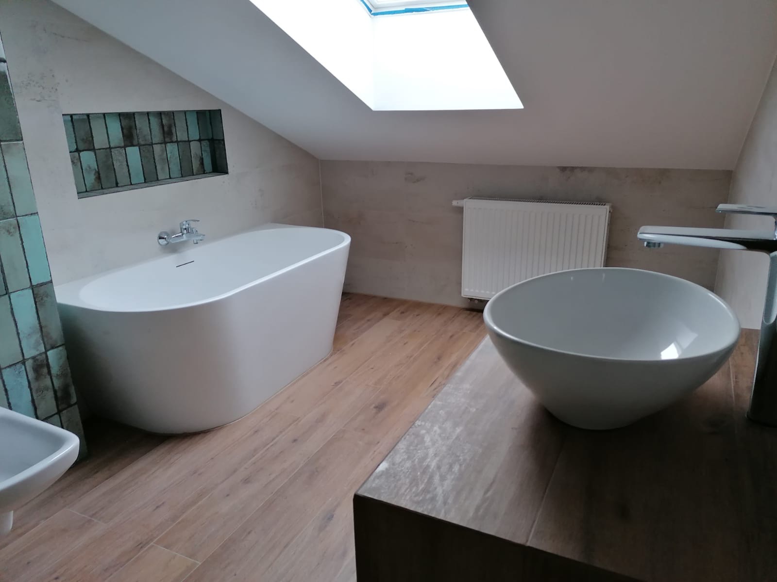 Nowoczesna łazienka na poddaszu ze skośnym sufitem, z wolnostojącą białą wanną, umywalką w kształcie misy na drewnianym blacie, grzejnikiem i oknem dachowym.