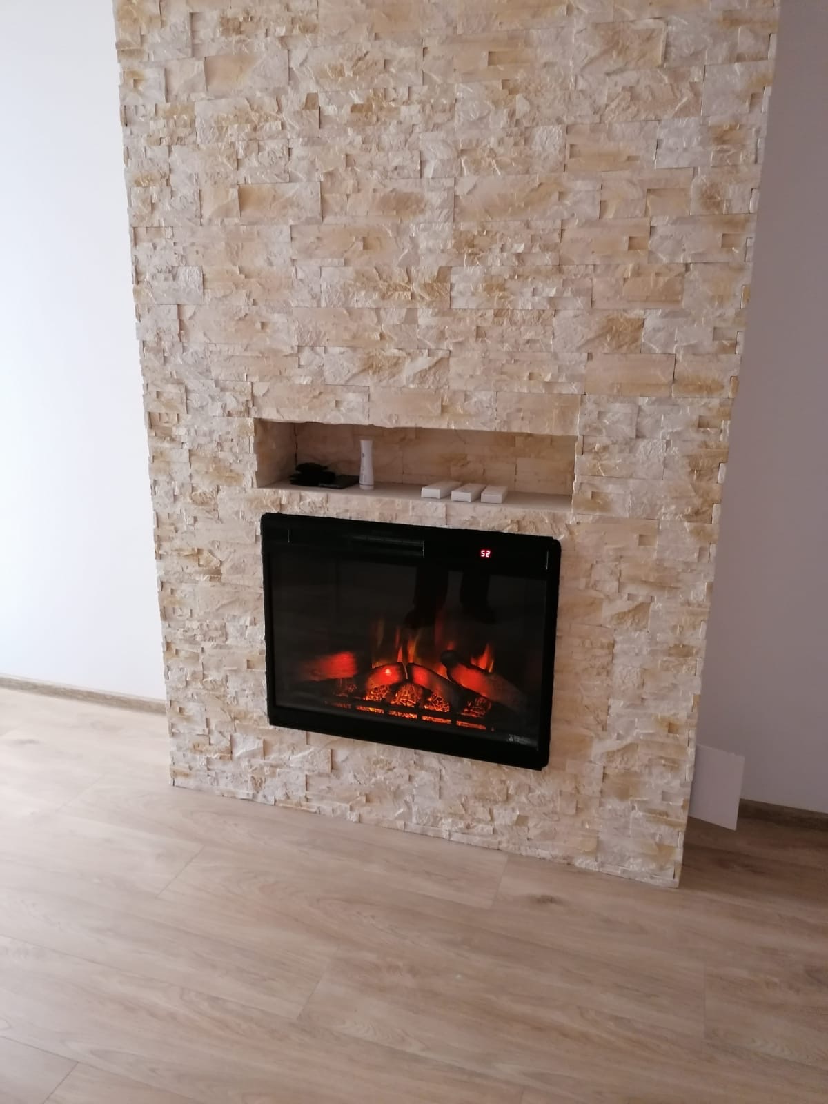 Ściana z dekoracyjnej, jasnej cegły z wnęką, w której umieszczony jest elektryczny kominek z efektem płomieni i imitacją drewna, na podłodze panele w odcieniach beżu.