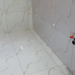 Świeżo wykafelkowana łazienka z białymi płytkami imitującymi marmur, widoczne fragmenty instalacji wodnej i surowy sufit z płyt kartonowo-gipsowych.