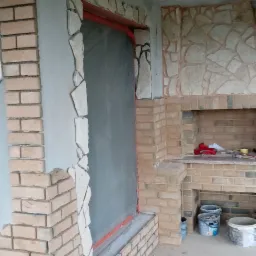 Murowanie kominka zewnętrznego z cegły klinkierowej i kamienia naturalnego, etap prac, widoczne wiadra z zaprawą i narzędzia.