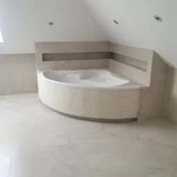 Wykończona łazienka na poddaszu ze skośnym sufitem. Uwagę przykuwa narożna wanna z mozaiką, otoczona płytkami imitującymi marmur. Minimalistyczne oświetlenie.