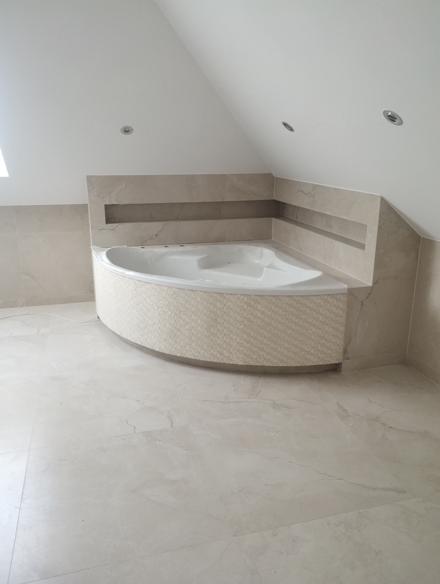 Wykończona łazienka na poddaszu ze skośnym sufitem. Uwagę przykuwa narożna wanna z mozaiką, otoczona płytkami imitującymi marmur. Minimalistyczne oświetlenie.