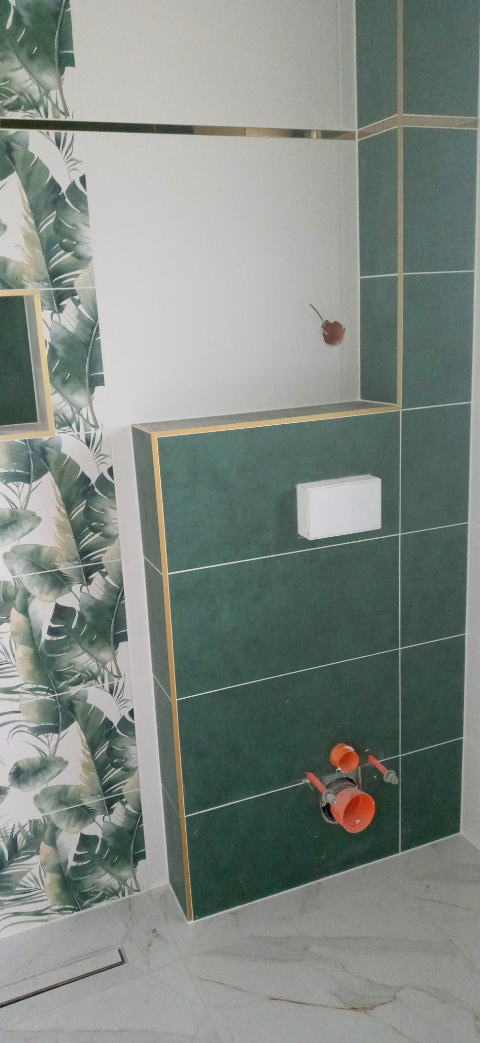 Fragment łazienki w trakcie wykańczania: zielone płytki z akcentami złota, wzorzysta ściana z motywem liści, widoczne elementy instalacji podtynkowej WC.